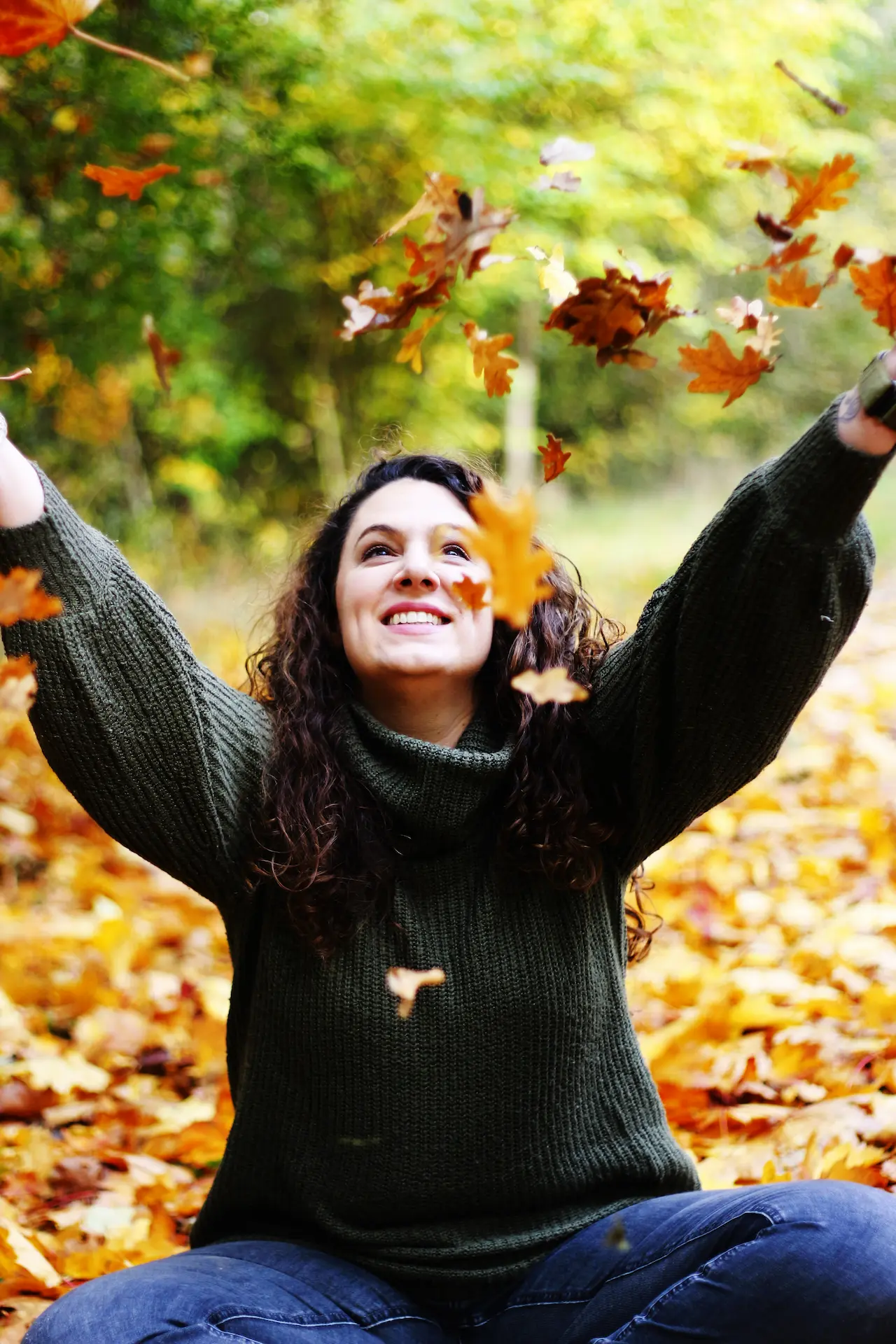 Coachin Liza aus Bremen-Huchting sitzt im herbstlichen Wald und wirft lächelnd bunte Blätter in die Luft. Symbol für Leichtigkeit und persönliches Coaching.